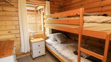 A wooden cabin room with bunk beds, a bedside table with a lamp, and a window with curtains.