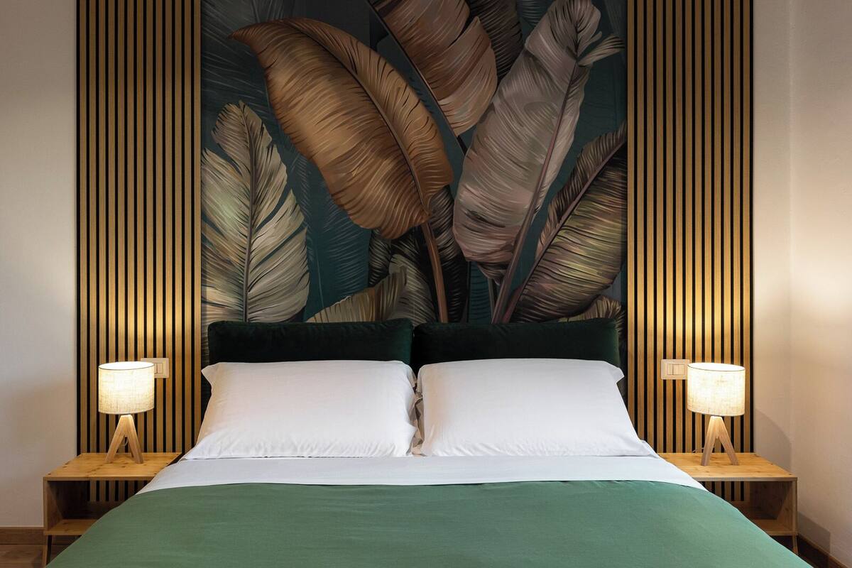 A bed with two pillows, flanked by two bedside tables with lamps, set against a wall with vertical wooden slats and a large tropical leaf mural.