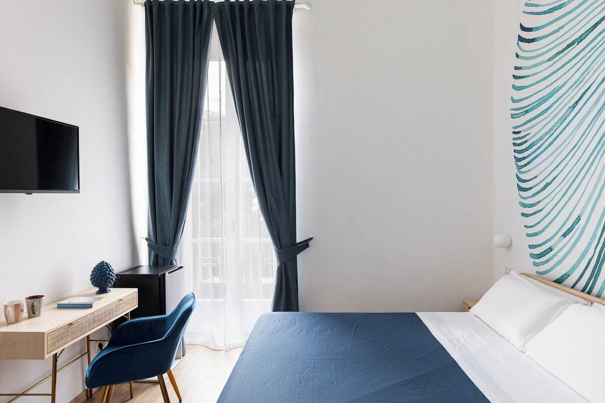 A hotel room with a bed, a desk with a chair, a television, and a window with curtains.