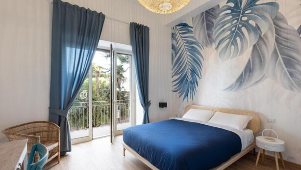 A bedroom with a bed, a chair, a small table, and a balcony with curtains.