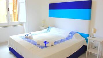 Una cama bien hecha con cabecera azul y blanca, una mesita de noche con lámpara, y una ventana con persianas.