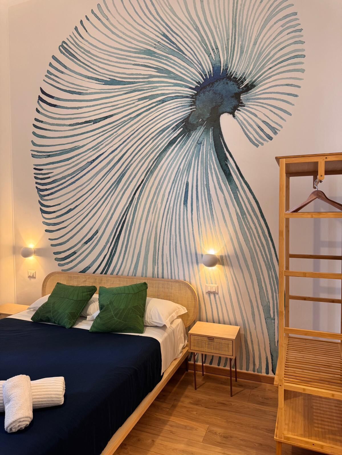 A bedroom with a large wall mural, a bed with green pillows, a wooden nightstand, and a wooden hanger stand.