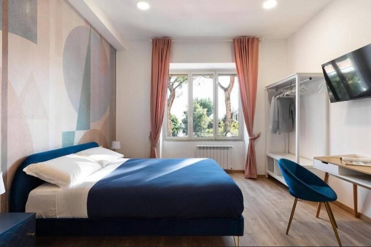 A modern hotel room with a bed, a desk, a chair, a TV, and a window with curtains.