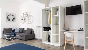 A modern living room with a grey sofa, a coffee table, a TV mounted on the wall, and a white shelving unit with clothes and a bicycle.