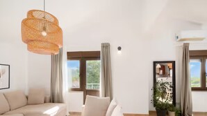 A modern living room with a beige sofa, a glass coffee table, and a hanging light fixture. There are large windows with curtains, a potted plant, and a view of greenery outside. - Villa Mima Pool Retreat (Lovreć)
