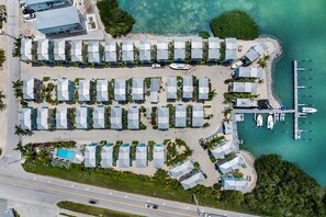 Oversigtsbillede af et resort med flere bungalows arrangeret i rækker langs en strand, en lysthavn med både og en vej, der løber parallelt med ejendommen.