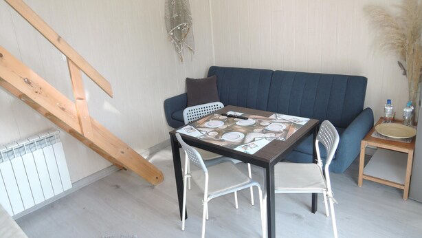 Un comedor con una mesa lista para cuatro personas, un sofá azul y una mesita auxiliar con un plato y botellas de agua.