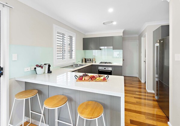 A modern kitchen with a central island, bar stools, a stainless steel refrigerator, and a window with blinds.