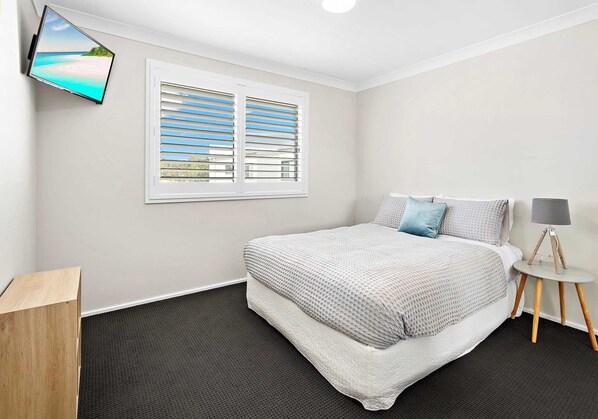 A bedroom with a bed, a window with blinds, a wall-mounted TV, a bedside table with a lamp, and a small wooden cabinet.
