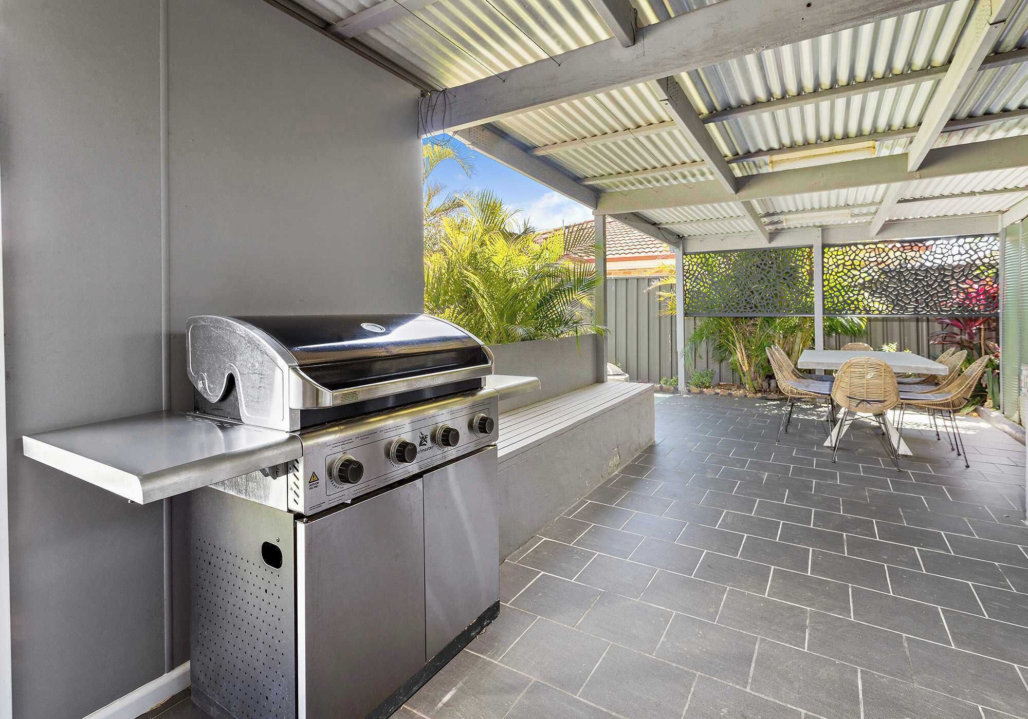 An outdoor kitchen with a stainless steel grill, a white countertop, and a tiled patio area with wicker chairs.