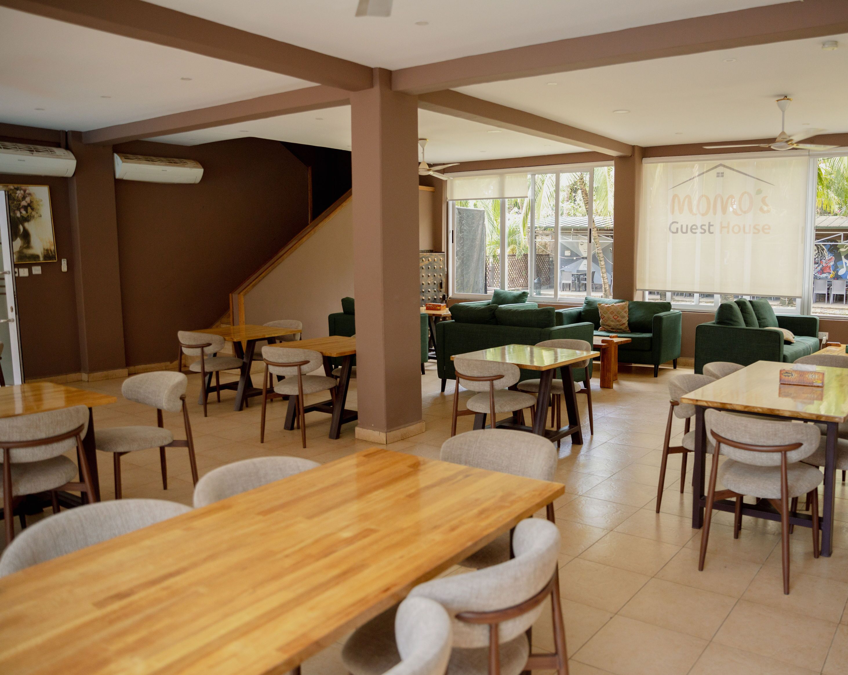 Un'ampia area interna con diversi tavoli e sedie in legno, un divano verde e una scala che conduce a un piano superiore.
