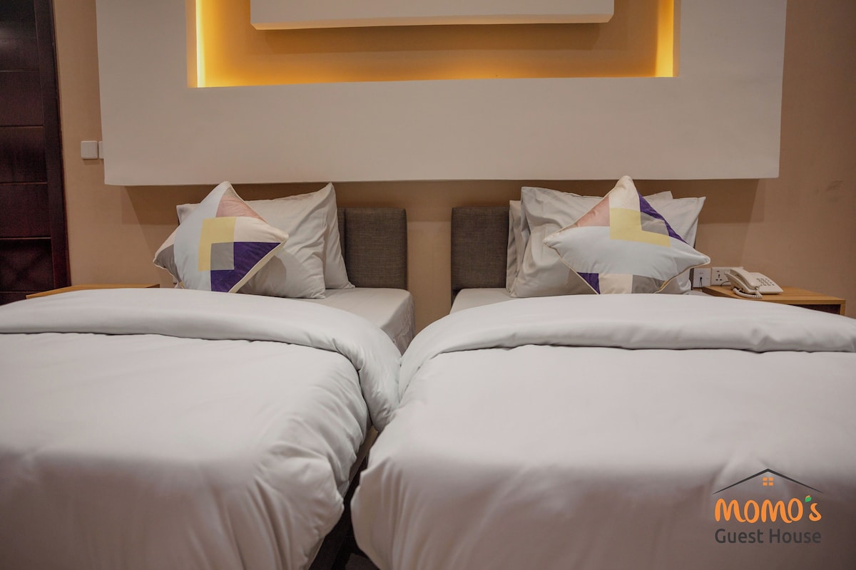 A hotel room with two beds, each with a pillow and a bedside table with a phone.