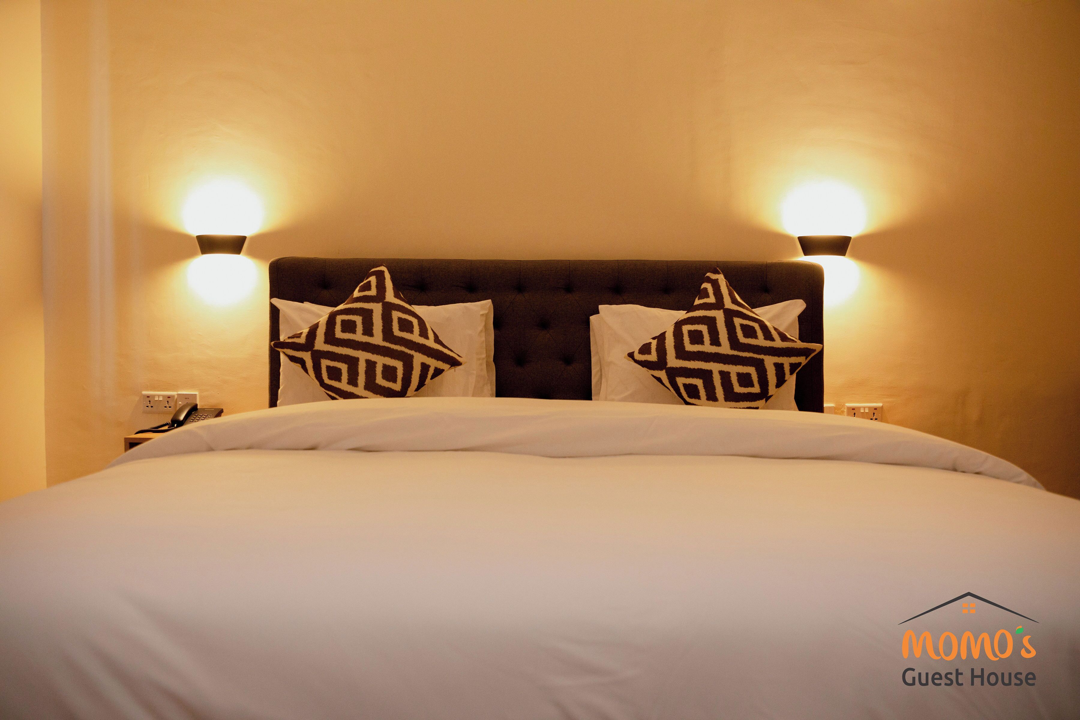 A neatly made bed with white linens and patterned pillows, a tufted headboard, and wall-mounted reading lamps.