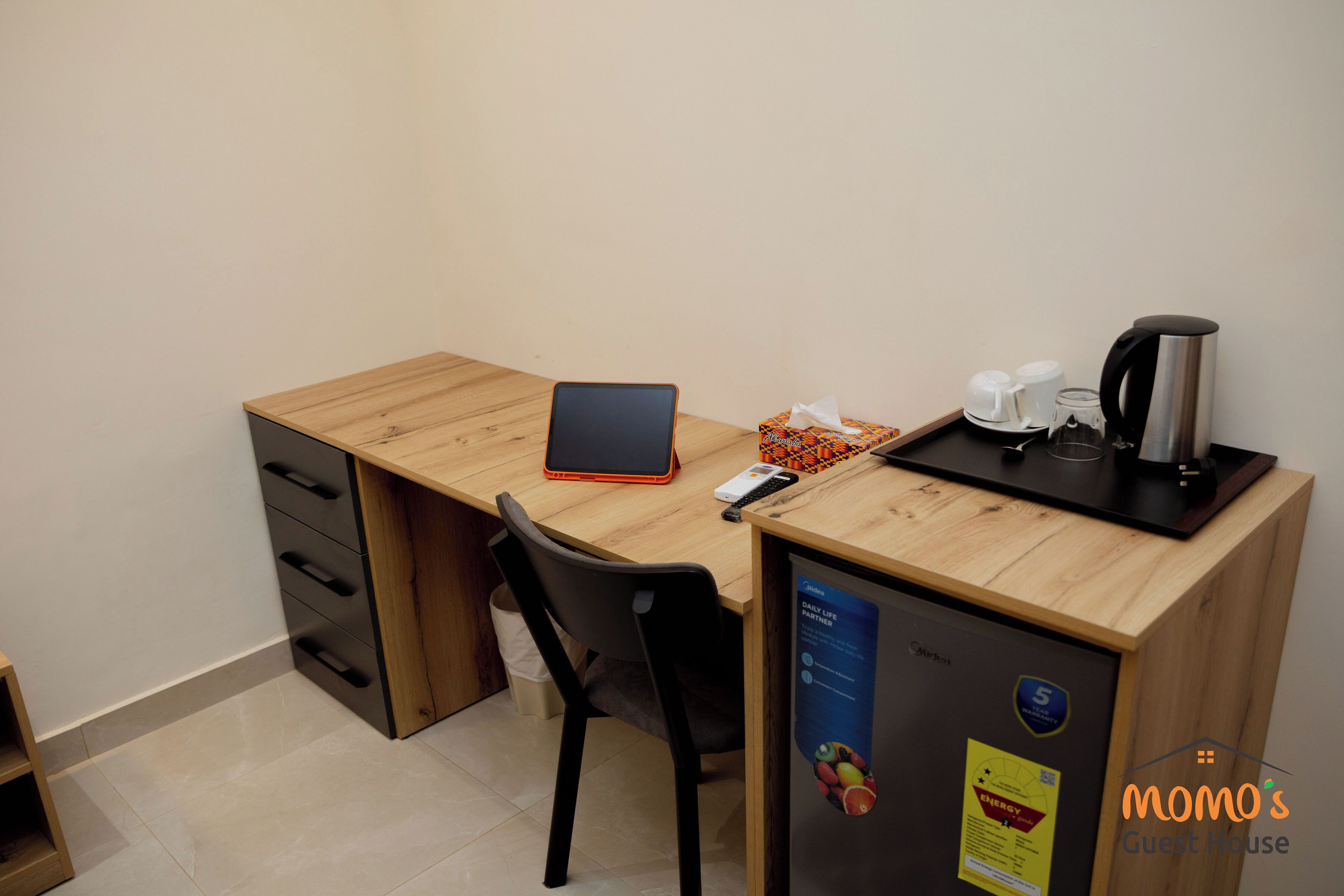 Ein Holztisch mit einem Laptop, einem Smartphone, einer Kaffeemaschine und einem kleinen Kühlschrank.