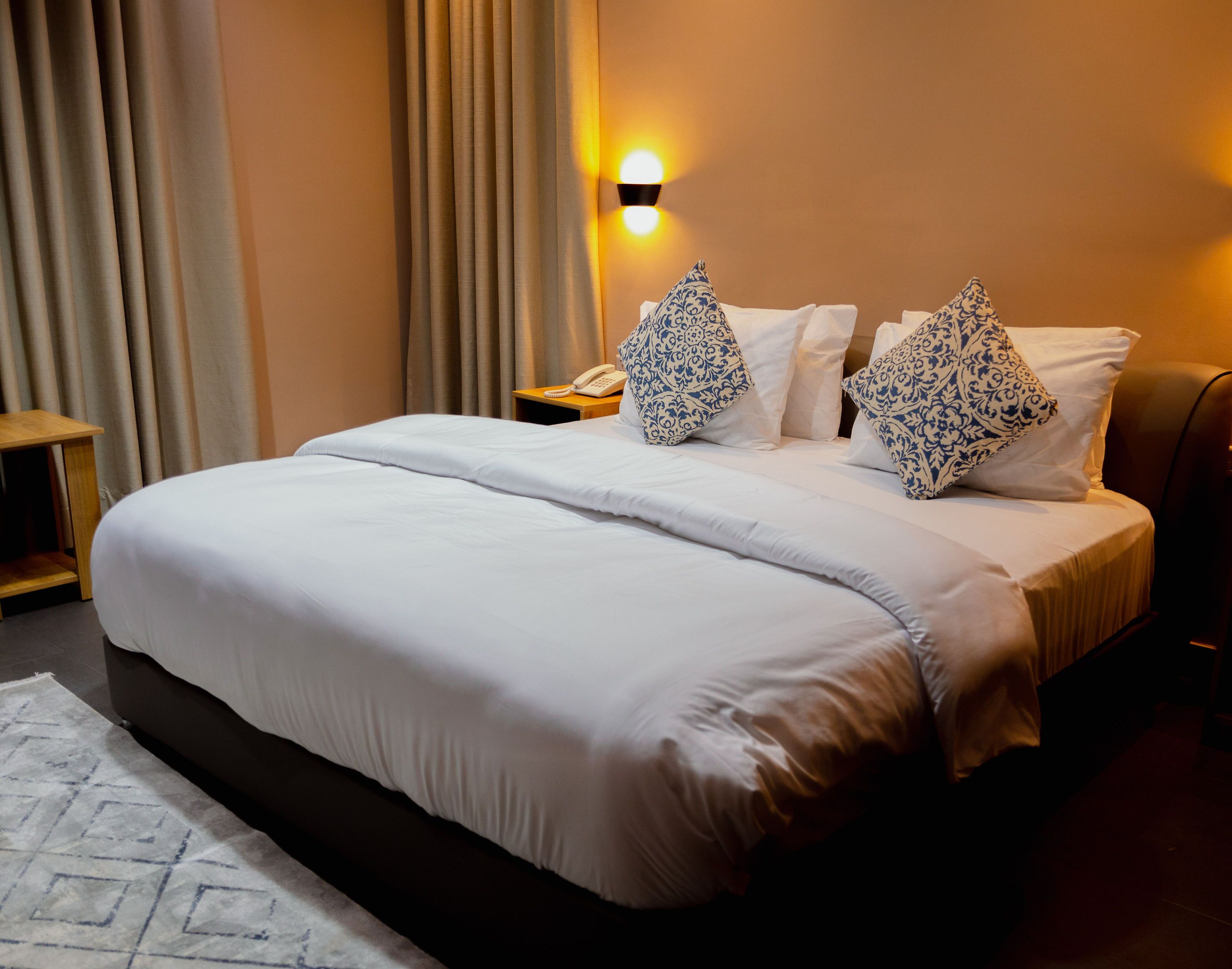 A neatly made bed with a white comforter and two decorative pillows, a bedside table with a telephone, and a wall-mounted light fixture.