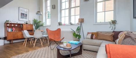 Un salon spacieux avec un haut plafond, de grandes fenêtres, un meuble en bois, un canapé, une table basse et un coin repas.