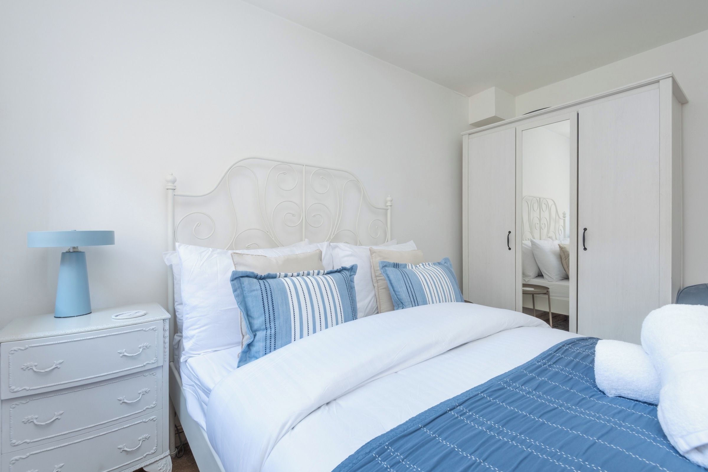 Un dormitorio con cama blanca, mesita de noche, lámpara, cómoda y armario.