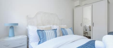 Un dormitorio con cama blanca, mesita de noche, lámpara, cómoda y armario.