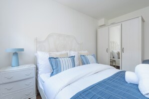 Ein Schlafzimmer mit einem weißen Bett, einem Nachttisch, einer Lampe, einer Kommode und einem Kleiderschrank.