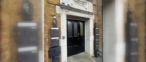A building entrance with a black door, a nameplate above the door, and mailboxes on either side.