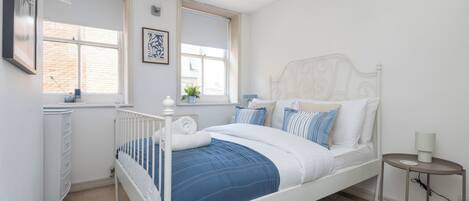 Un dormitorio con una cama con dosel blanco, una mesita de noche pequeña con una lámpara y una ventana con vista a un edificio de ladrillo.