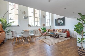 A spacious living area with a dining table, chairs, a sofa, and a coffee table. There are large windows providing natural light and a view of the outside. - Sunlit 3BDR Loft Near Tooting Broadway (Greater London)