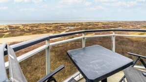 Un balcón con mesa y sillas que da a un paisaje costero con vegetación escasa y una vista distante del mar.