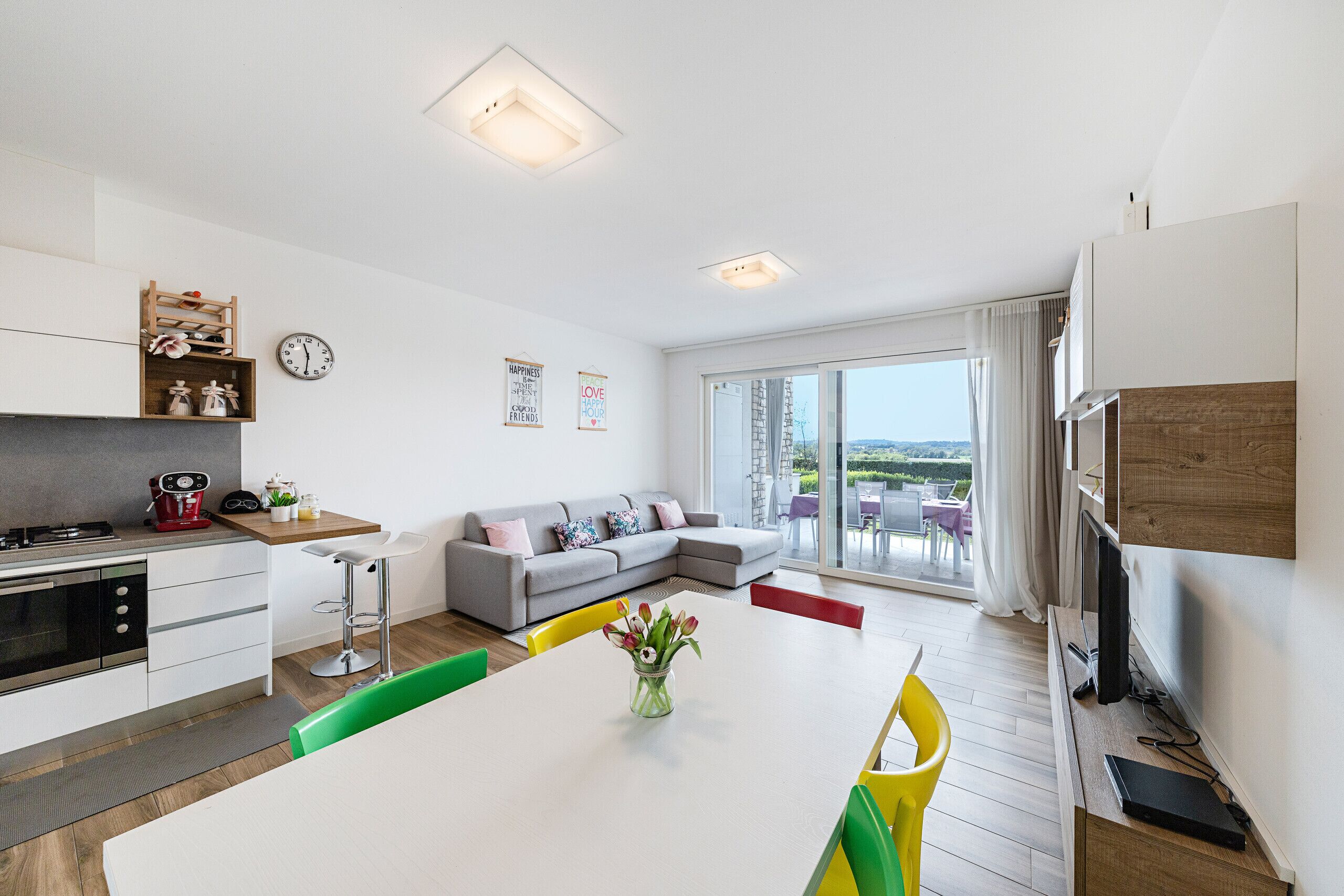 A modern kitchen with a dining area, a grey sofa, and a large window with a view.