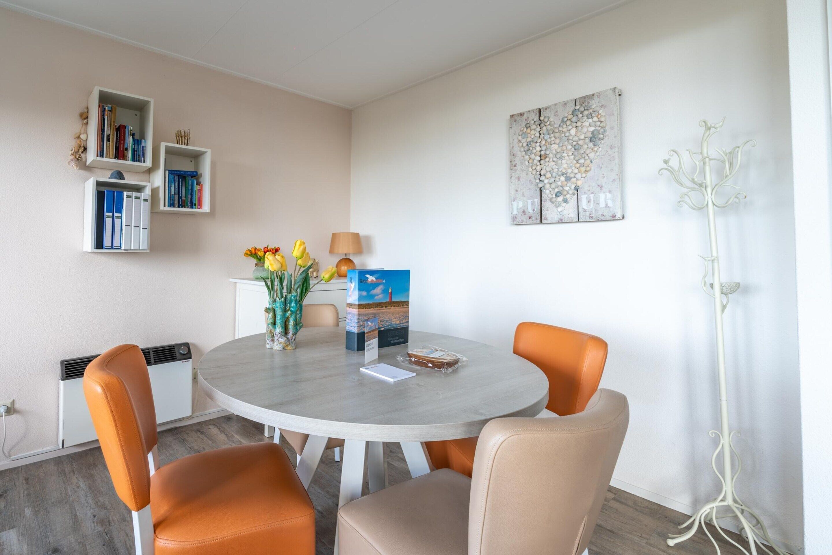 A dining area with a round table, four chairs, a vase with flowers, a lamp, and bookshelves.