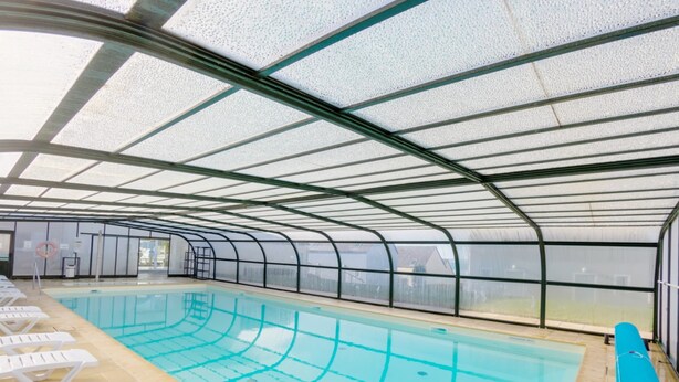Une piscine intérieure à l’eau cristalline, entourée de chaises longues blanches et surmontée d’un plafond en verre.