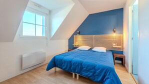 Une chambre avec un plafond en pente, une grande fenêtre, un lit recouvert d’une courtepointe bleue et une tête de lit en bois agrémentée de lampes fixées au mur.