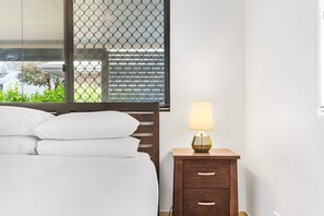 Ein ordentlich bezogenes Bett mit weißen Leinen, ein Nachttisch aus Holz mit einer Lampe und ein Fenster mit Jalousien.