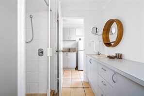 Ein modernes Badezimmer mit weißen Fliesen, einer Dusche, einem Waschbecken mit Holzspiegel und einer Küche im Hintergrund.