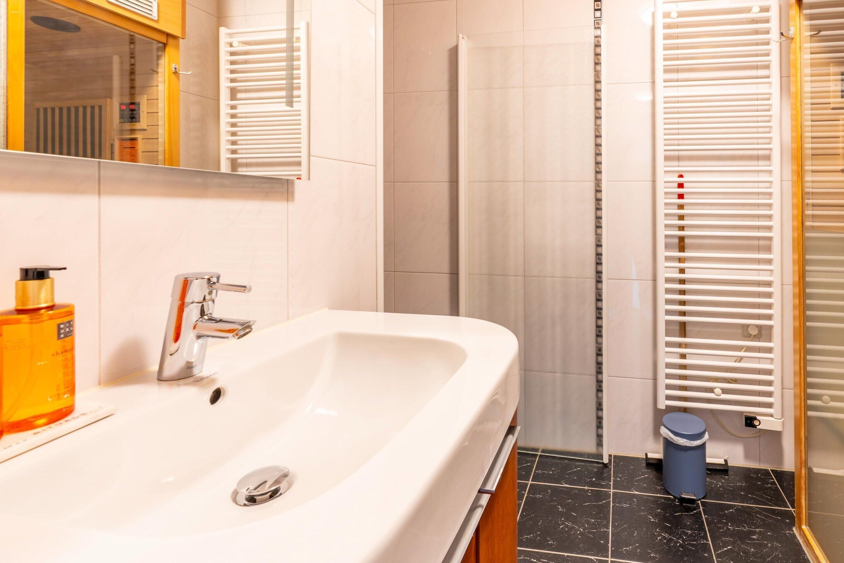 A bathroom with a white sink, a mirror, a bottle of soap, and a blue trash bin.