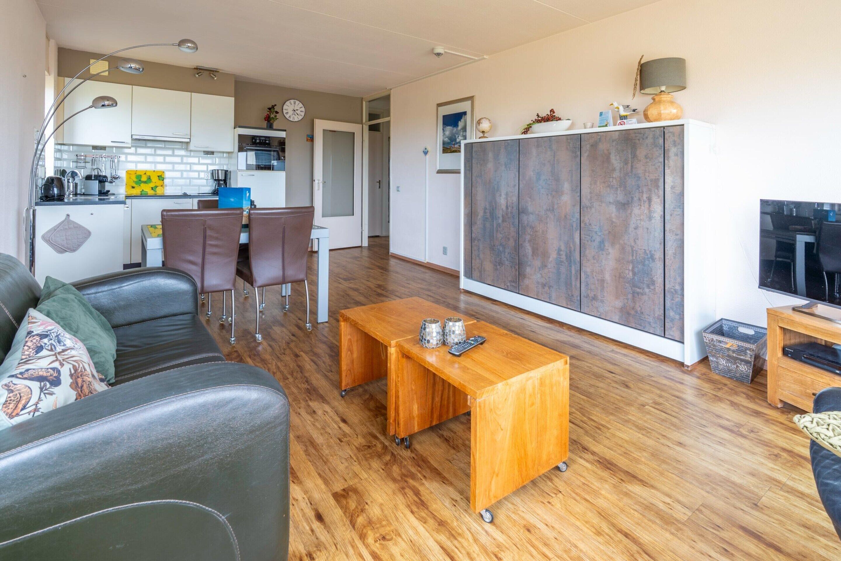 A modern living room with a sofa, coffee table, and a TV. A kitchen is visible in the background with a dining area.