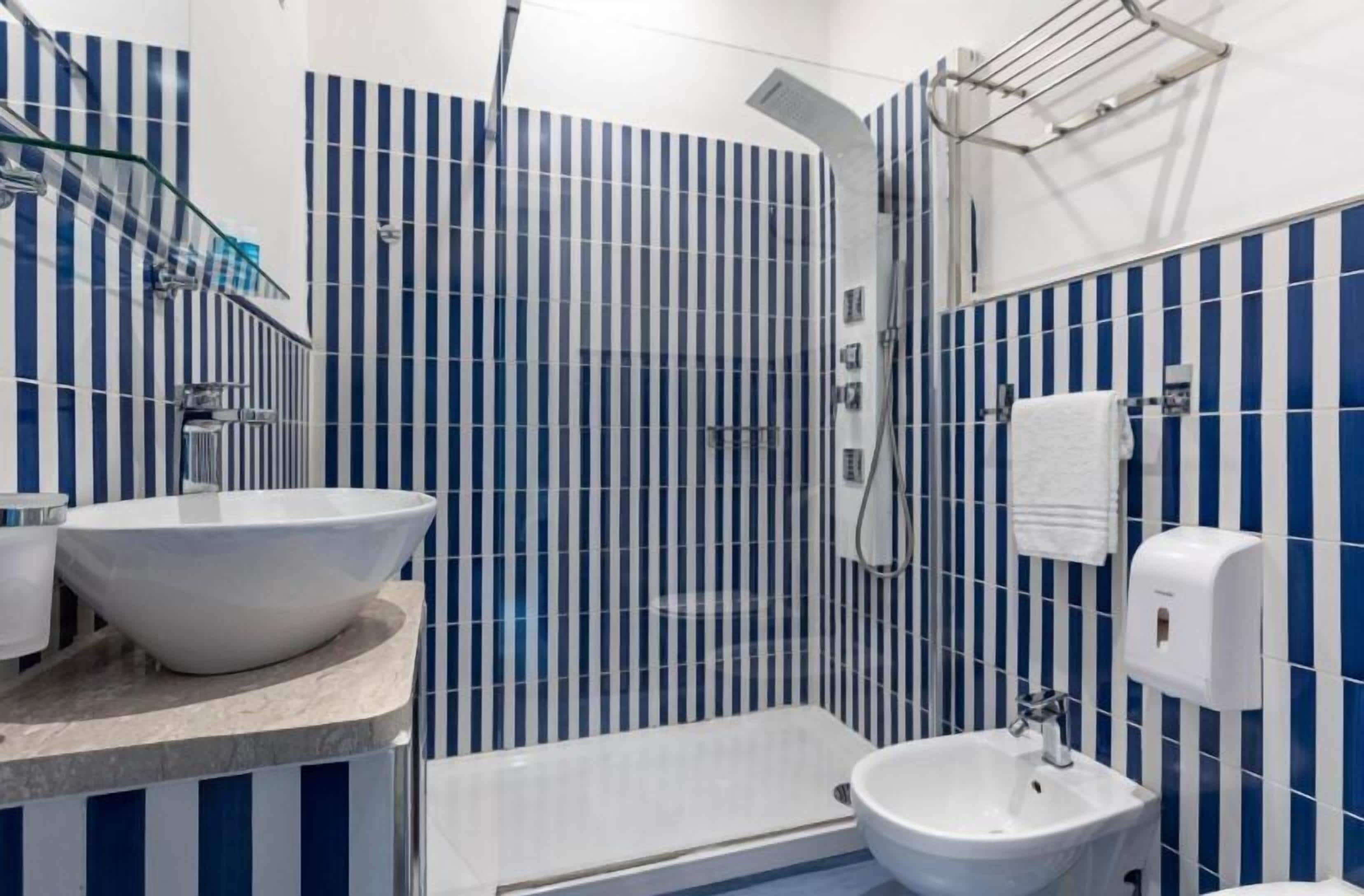 Ein Badezimmer mit einem großen weißen Waschbecken, einem kleineren Waschbecken, einer Duschkabine aus Glas und einem Handtuchspender.