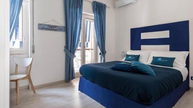 A bedroom with a large bed, blue curtains, a white chair, and a wall-mounted air conditioner.