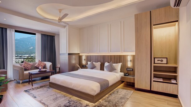 A modern hotel bedroom with a large bed, bedside tables, a nightstand, a wardrobe, and a view of mountains through the window. - Hotel Urban chant (Rishikesh)