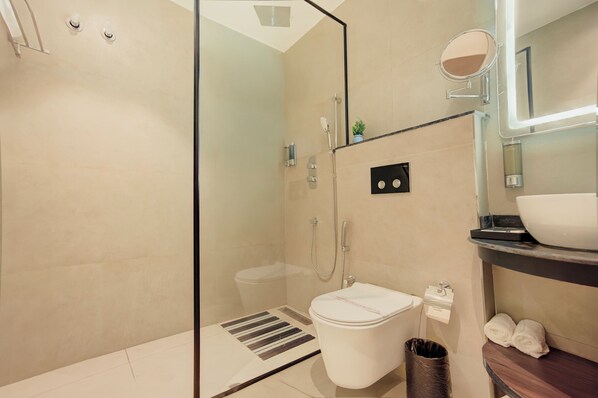 A modern bathroom with a glass shower enclosure, a white toilet, a vanity with a mirror, and a trash can. - Hotel Urban chant (Rishikesh)