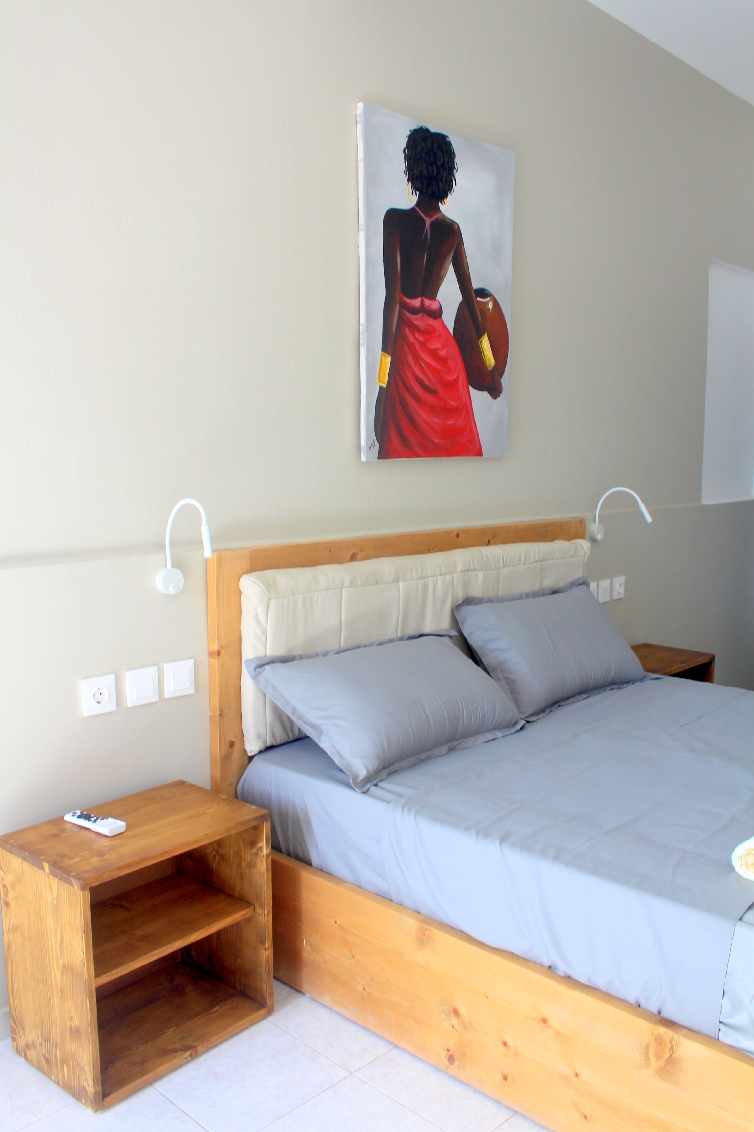 A bedroom with a wooden bed frame, grey bedding, a small wooden nightstand, and a wall-mounted light fixture.