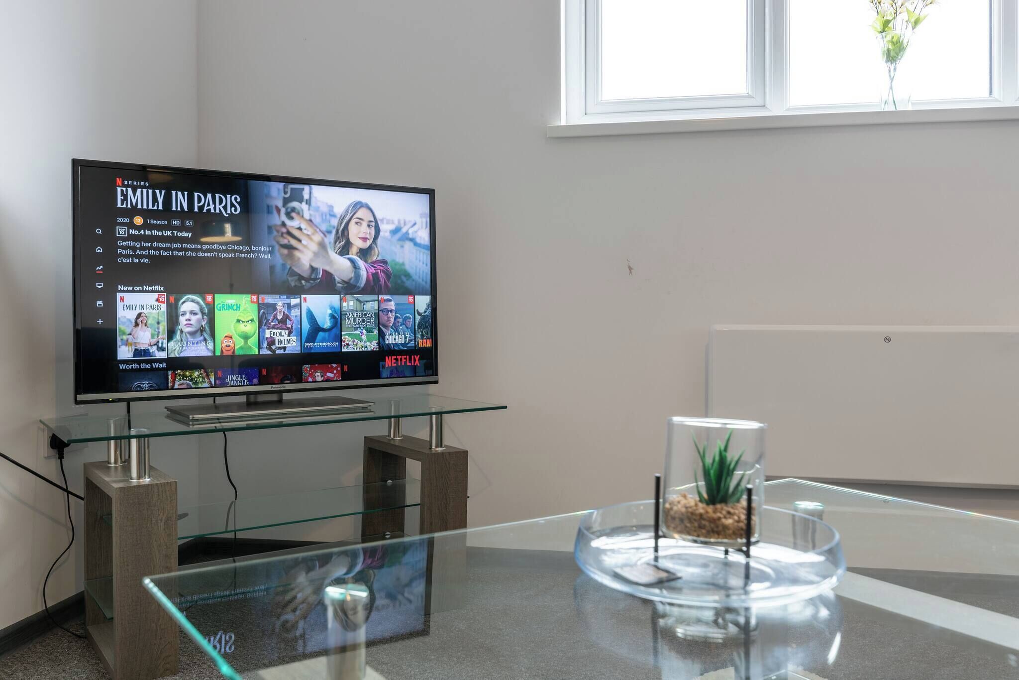 Quarto de hotel moderno com uma mesa de centro de vidro, uma TV de tela plana exibindo a interface da Netflix e vista para uma janela com uma planta.