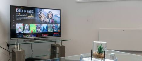 Een moderne hotelkamer met een glazen salontafel, een flatscreen televisie waarop een Netflix-interface te zien is, en een uitzicht op een raam met een plant.