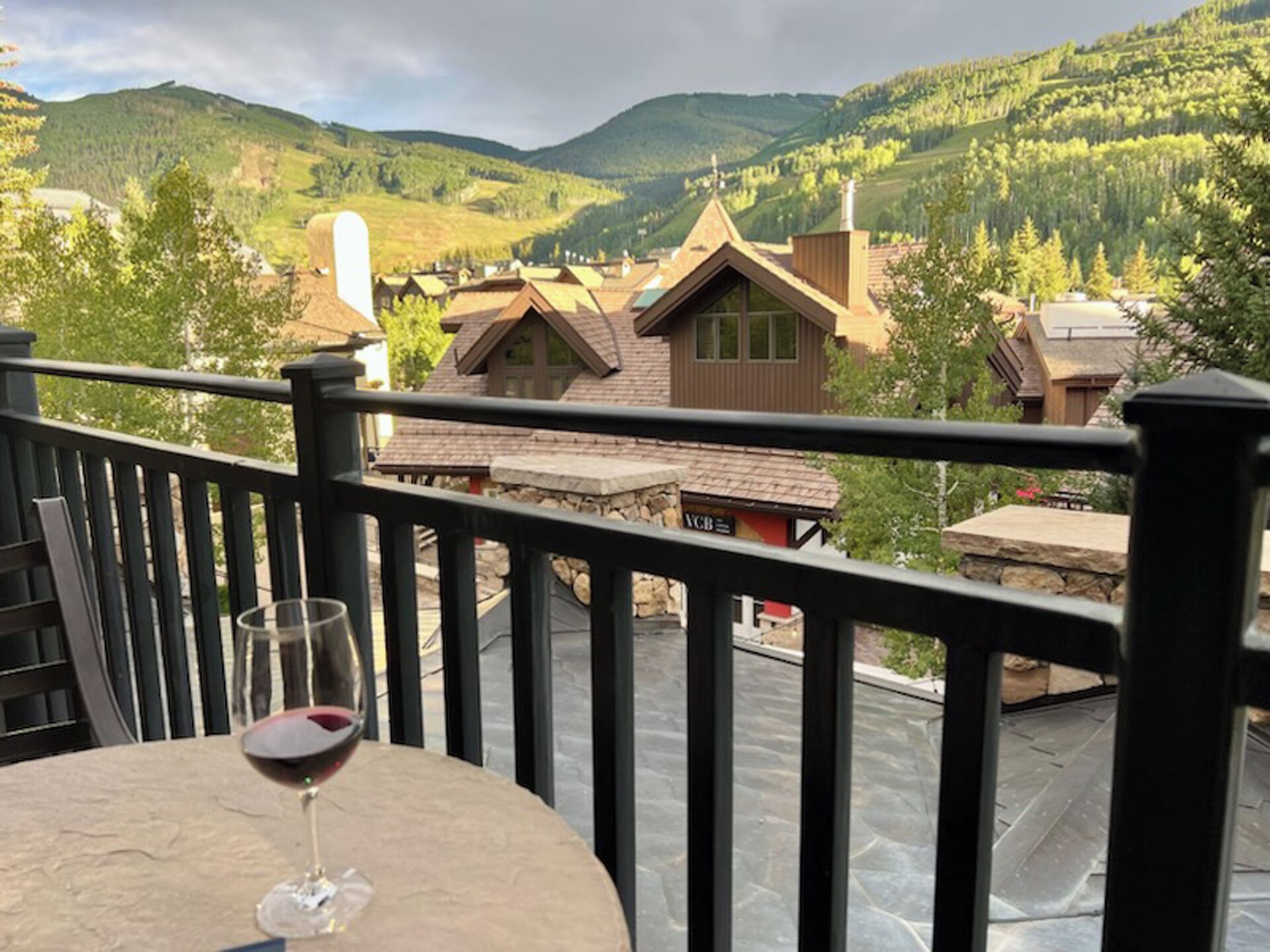 Un balcon avec une table et un verre de vin rouge, donnant sur un village aux maisons traditionnelles et un paysage montagneux.