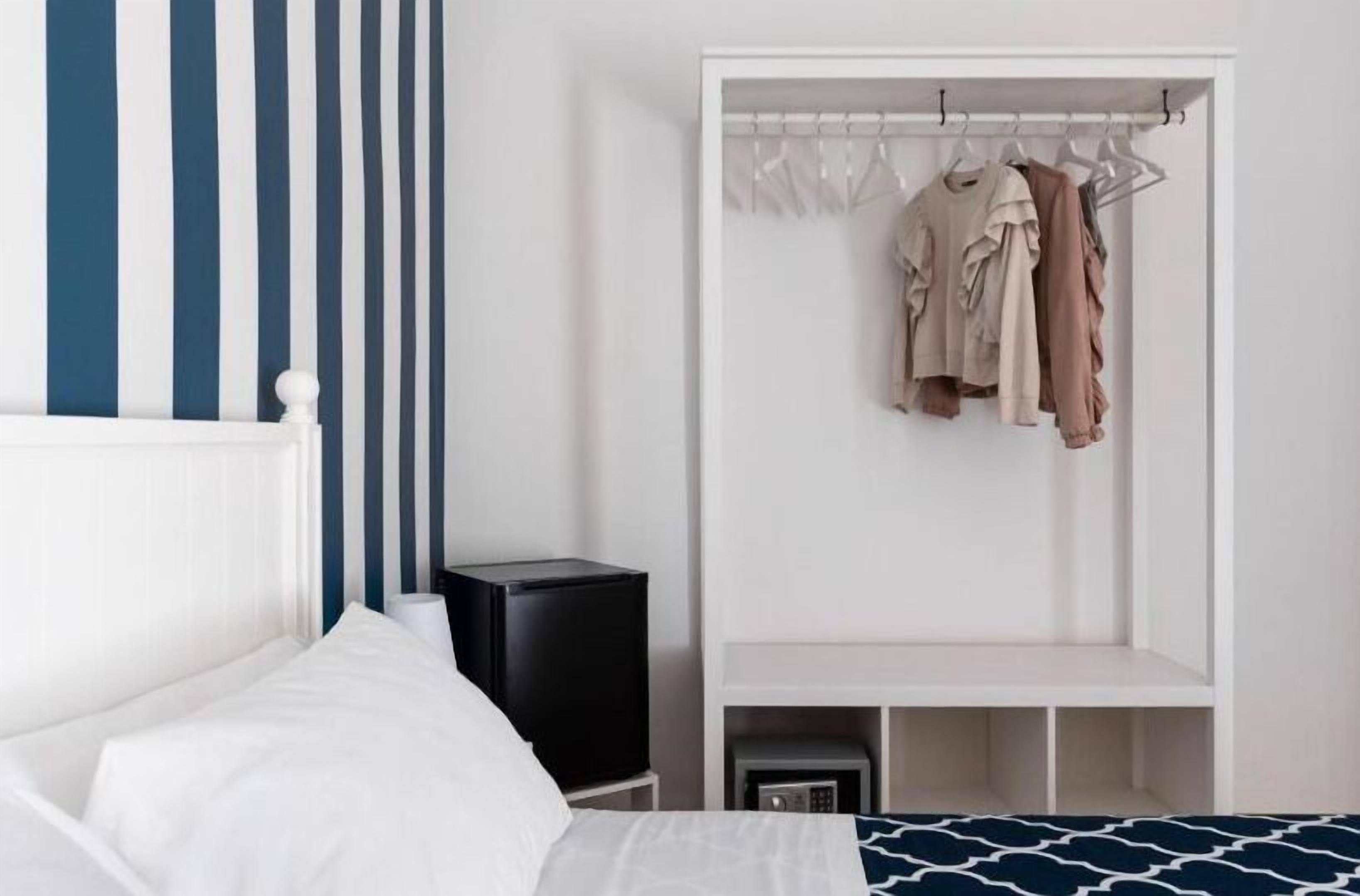 A hotel room with a bed, a white wardrobe with hanging clothes, a black speaker, and a small bedside table.