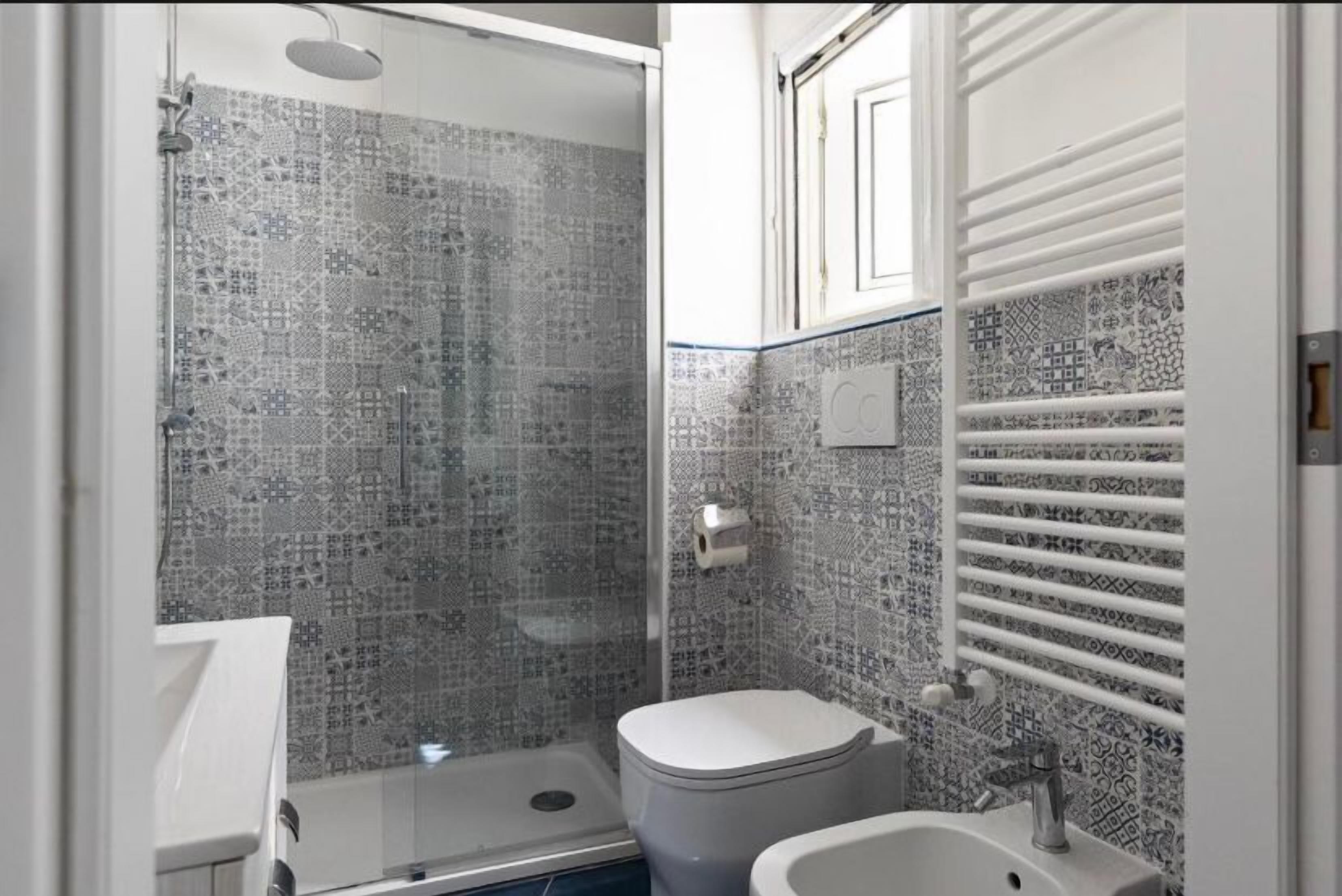 Un baño con ducha, inodoro y lavamanos, con paredes de azulejos estampados y un tocador blanco.