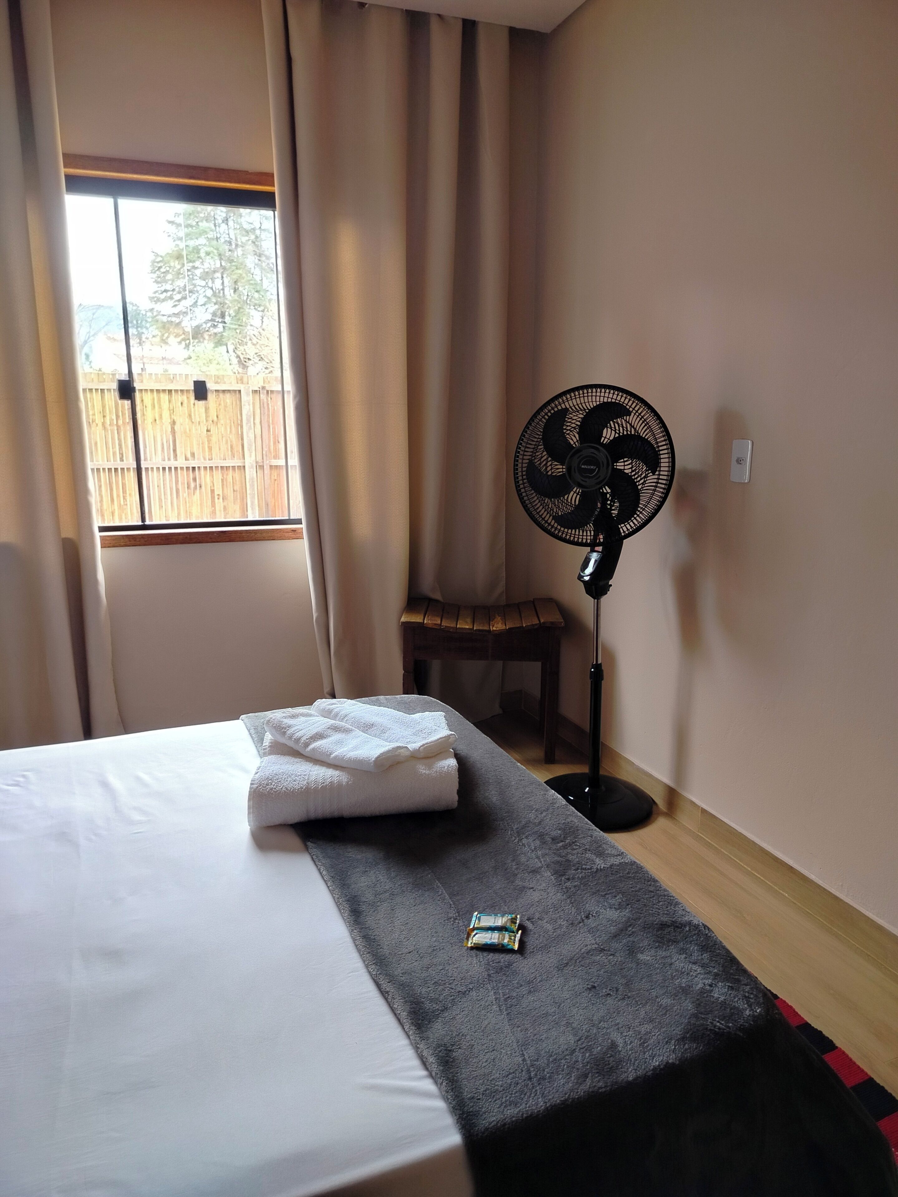 Habitació d'hotel amb un llit, un ventilador, una taula petita i una finestra amb cortines.