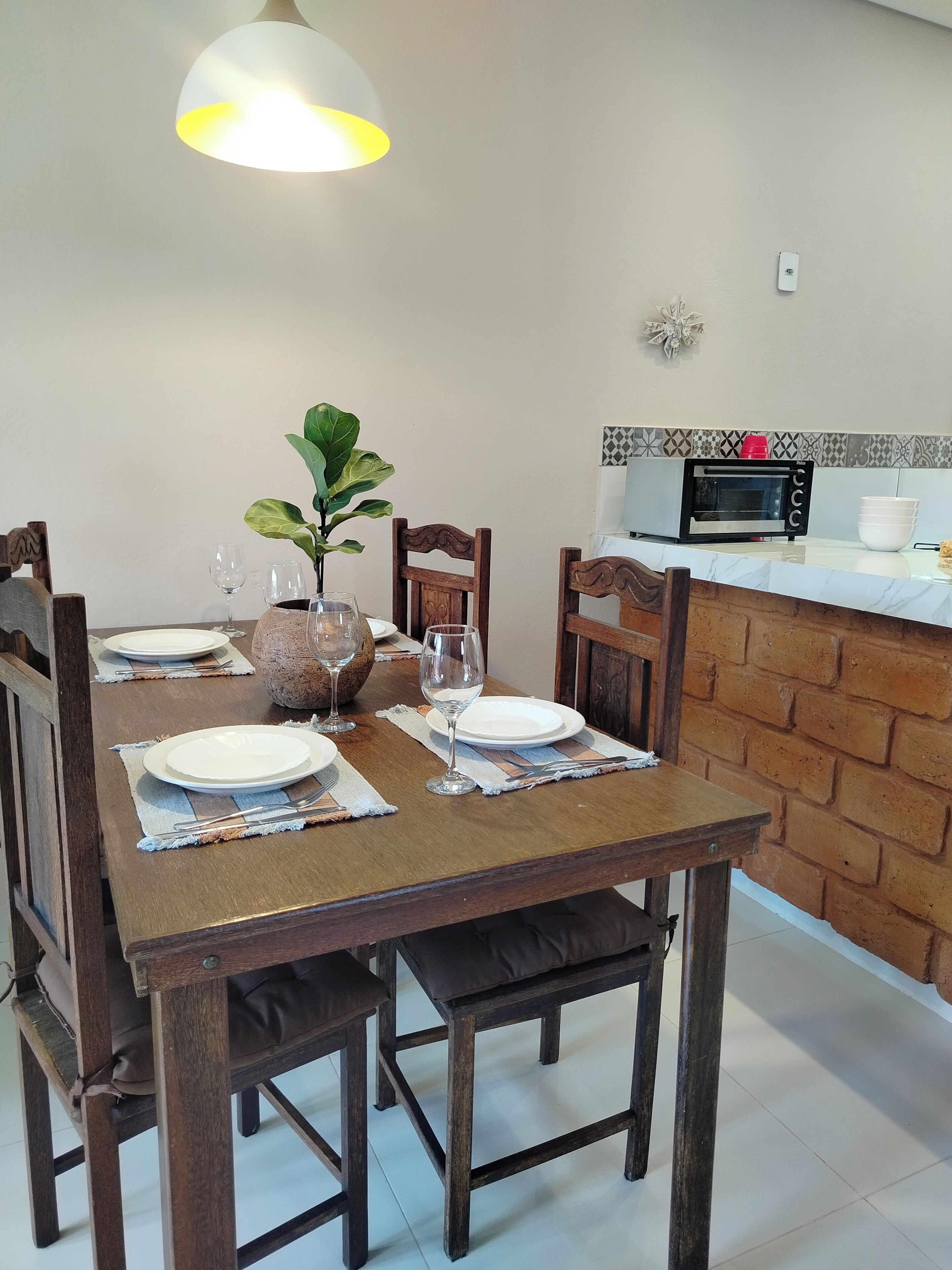 Una mesa de comedor dispuesta con platos, vasos y un florero con una planta, en una habitación con pared de ladrillo y un microondas.