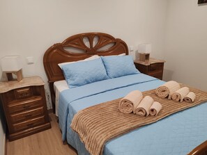 Un dormitorio con cabecera de madera, cama con ropa de cama azul, dos mesitas de noche con lámparas y un aparador de madera.