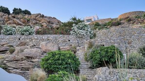 A stone wall with a garden in front, a building with a sloped roof, and a clear sky. - Aloft Suites Mykonos Island (Mykonos)