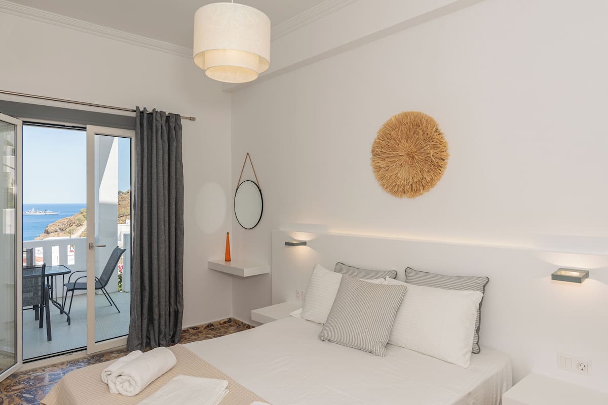 A bedroom with a bed, a bedside table, a lamp, a round wall decoration, a window with curtains, and a view of the sea.